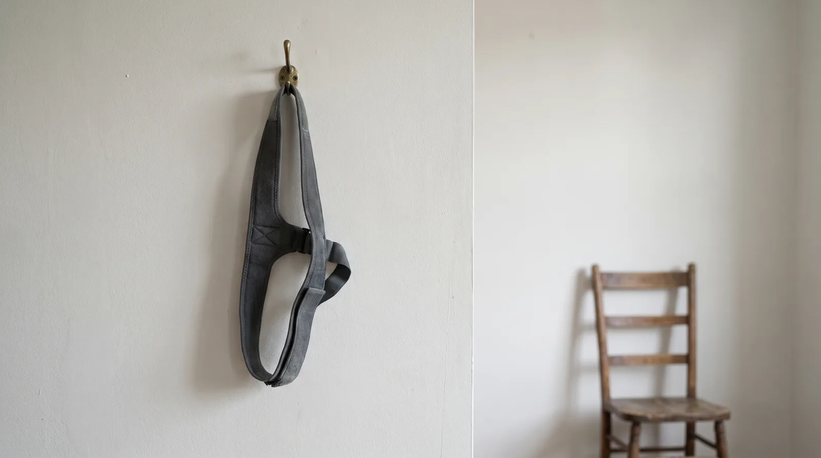 A posture-correcting shoulder brace hanging on a hook on a plain wall, with an empty chair in the background, soft neutral lighting
